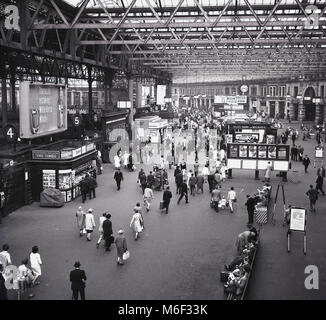 Années 1960, 09.15h, et un tableau historique du grand hall de la gare de Londres Waterloo, Lambeth, London, England, UK, montrant la structure de l'intérieur, les gens et les panneaux publicitaires pour les journaux nationaux tels que le Times et des boissons comme le whisky du Cheval Blanc. Banque D'Images