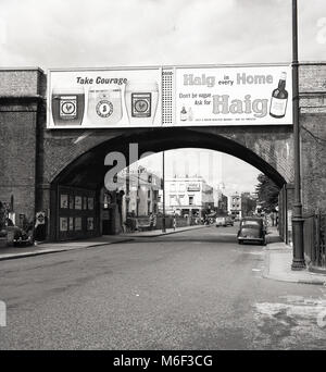 60, tableau historique d'un quartier calme de Kentish Town Road, London, NW1, montrant de grands panneaux publicitaires pour les grandes marques de boissons de la journée, le courage de la bière et whisky Haig sur un pont de chemin de fer. Au loin, un signe pour une autre bière bien connue de l'époque, la "double Diamond' par Ind Coope au bras de Moreton pub. Banque D'Images