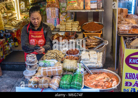 Une femme regarde son smartphone lors de l'attente pour les clients de son street food sur un sentier à Hanoi, Vietnam Banque D'Images