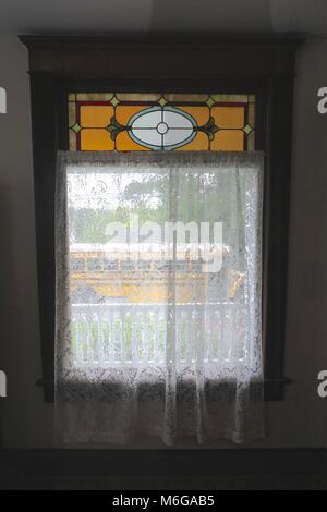 Fenêtre avec des rideaux de dentelle blanche, un petit vitrail au-dessus, donnant sur un porche blanc et un autobus scolaire. Banque D'Images