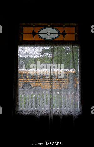 Fenêtre avec des rideaux de dentelle blanche, un petit vitrail au-dessus, donnant sur un porche blanc et un autobus scolaire. Banque D'Images