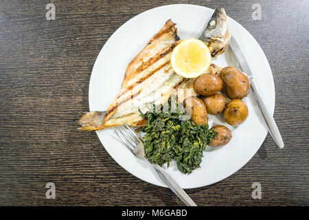 Loup de mer grillé servi avec du citron, pommes de terre au four et les épinards on white plate Banque D'Images