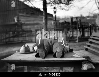 Un homme dernier repose sur une table d'extérieur avec un cigare dans la bouche et l'écriture sur ses chaussures, ca. 1910. Banque D'Images