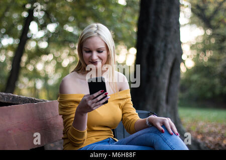 Très jolie jeune femme en utilisant smartphone outdoors dans un parc, assis sur un banc. Banque D'Images