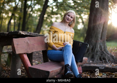 Jeune femme blonde assise sur un banc, écouter de la musique sur son smartphone à l'extérieur dans un parc. Heureux, se détendre. Banque D'Images