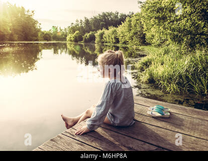 Adorable petite fille assise sur le pont de bois et à la recherche sur la rivière et le soleil Banque D'Images