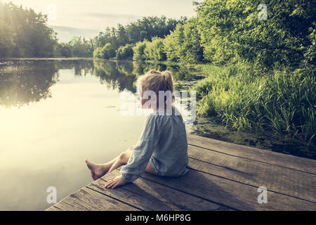 Petite fille solitaire rêvant sur un quai près de petit lac sur chaude journée d'été Banque D'Images