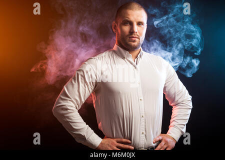 Beau jeune homme sportif en chemise blanche est souriant et en toute confiance, regarde la caméra en face de rouge et bleu fumée d'une lingette sur une isol noir Banque D'Images