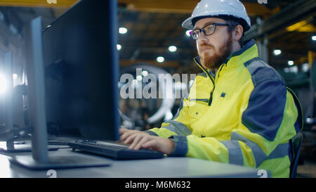 Ingénieur industriel travaille à l'espace de travail sur un ordinateur personnel. Il porte un casque et veste de sécurité et travaille à l'atelier principal de la Lourde Corp. Banque D'Images