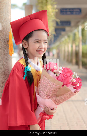 Petite fille asiatique en rouge robe du diplômé sourire avec bonheur avec bouquet de fleurs à la main Banque D'Images