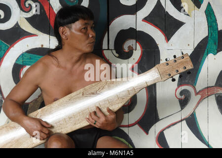 L'homme natif jouant la musique traditionnelle sur une bouilloire sape dans l'Orang Ulu longue maison, avant d'arbre de vie murale dans le village culturel de Sarawak Banque D'Images