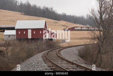 Les voies de chemin de fer leur courbe façon par un paysage rural de granges rouge sombre au cours d'une saison d'hiver. Banque D'Images