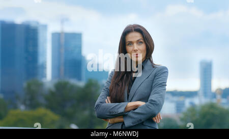 Femme d'affaires réussie traverse les bras et sourit en arrière-plan des gratte-ciel et un centre d'affaires de la grande ville est visible. Banque D'Images