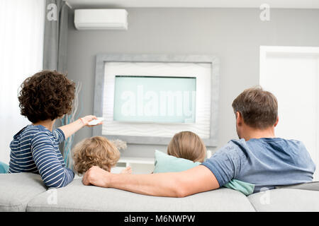 Retour Voir portrait de famille heureuse avec deux enfants de regarder la télévision assis sur un canapé dans la salle de séjour et d'allumer la télévision moderne avec télécommande contr Banque D'Images