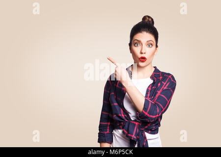 Jeune fille avec surprise et style décontracté cheveux chignon pointant son doigt sur le côté, montrant quelque chose sur le mur blanc beige avec copie espace pour votre Banque D'Images