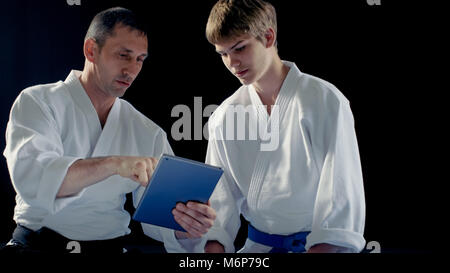 Maître d'Arts Martiaux portant des Hakamas enseigne aux jeunes élèves Aikido technique avec l'aide de l'ordinateur tablette. Shot isolé sur fond noir. Banque D'Images