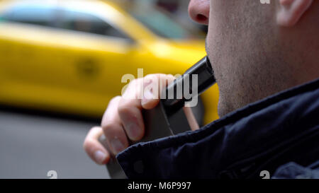 Jeune homme millénaire hipster fumeurs vape e-cigarette à l'extérieur sur la rue de la ville de New York pendant la journée. Porter veste bleue, jaune taxi passe par Banque D'Images