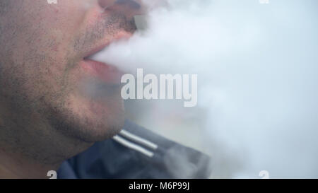 Jeune homme blanc soufflé vape la fumée dans la bouche à l'extérieur sur la rue de la ville. Hipster millénaire urbain habitude addictive de dispositifs e-cigarette Banque D'Images