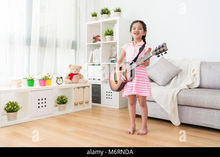 Joyeuse petite fille jouant de la guitare en bois en face du canapé. Sourire gaiement à l'appareil photo. Sur une vue de côté. Elle est la pratique de la guitare moins Banque D'Images