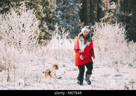 Belle Jeune Fille Pretty Caucasian Woman Dressed In White Hat et Red Jacket jouant avec chiot de race mélangée chien dans la forêt enneigée en hiver 24. Banque D'Images