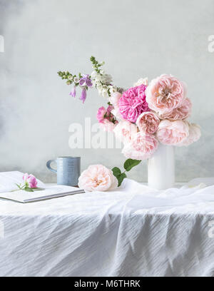 Still Life, piscine, vue de face de table couverte de draps gris pâle, avec vase blanc rose de David Austin Roses & digitales, mug bleu pâle, à livre ouvert Banque D'Images