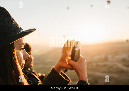 Vue latérale du charming brunette girl in hat prend photo sur smartphone pendant les vols de lots de montgolfières sur chaude journée ensoleillée. Ta femme brune Banque D'Images