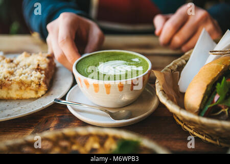 Close-up man's hand holding mug de thé vert avec de beaux modèle dans la forme de mousse blanche à côté de dessert et de sandwich Banque D'Images