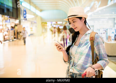 Billet femme debout avec les bagages à l'aéroport. en utilisant son smartphone SMS message autour de la boutique duty free dans l'aéroport international. Banque D'Images