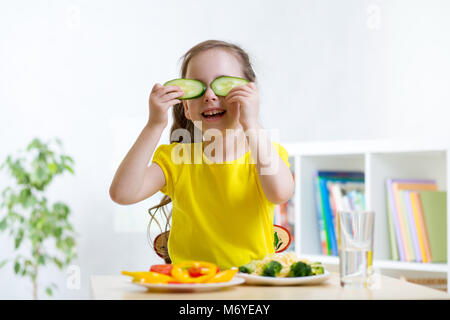 Happy little girl having fun with food légumes tandis que le dîner. Elle détient les concombres sous ses yeux comme dans les verres. Banque D'Images