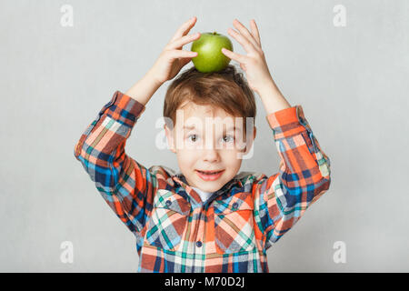 Smiling boy holding a green apple sur sa tête, dans une chemise à carreaux, sur fond gris Banque D'Images