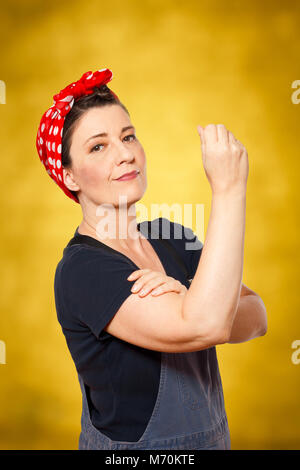 Sûr de femme d'âge moyen avec salopette retrousser sa manche, l'espace texte, rendre hommage à l'icône de l'ouvrier américain à riveter Rosie Banque D'Images