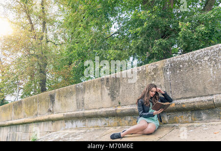 Belle fille brune de la lecture d'un vieux livre, extérieur, sur un mur en pierres, sur une journée ensoleillée. Banque D'Images