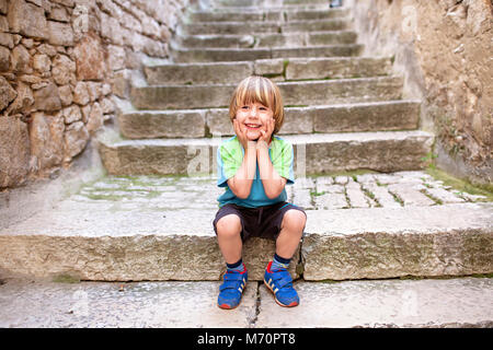 Cute smiling blonde petit garçon assis sur de vieux escaliers pavés avec les mains sur les joues, dans le centre historique de Rovinj Banque D'Images