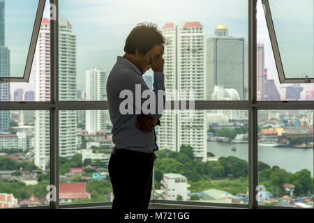 L'homme déprimé face permanent dans les mains sur l'intérieur du gratte-ciel avec faible éclairage à côté de l'arrière-plan de windows sur le paysage urbain, dramatique Banque D'Images