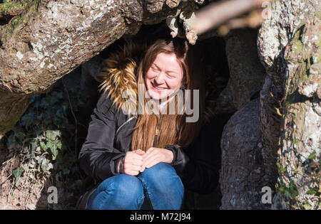 Jolie jeune femme assise à l'extérieur sous un arbre portant un manteau, par une froide journée d'hiver avec du soleil sur son visage. Banque D'Images