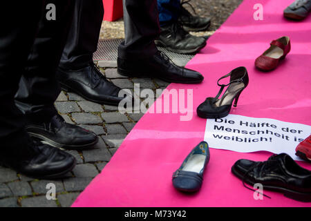 08 mars 2018, l'Allemagne, Stuttgart : hommes debout par un tapis, qui est couverte d'affiches. Le Conseil d'État sont des femmes à l'aide de cette campagne de lutte pour l'égalité entre les hommes et les femmes : Un tapis rose avec une rangée de paires de chaussures chaussures unique composé de des hommes et des femmes. Photo : Sina Schuldt/dpa Banque D'Images