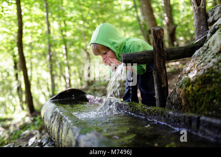 Jeune garçon blond l'eau potable de creux des mains de l'auge rustique dans une forêt Banque D'Images