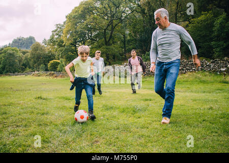 Trois génération de la famille jouent au football ensemble dans un champ. Il y a deux garçons, leur père et leur grand-père. Banque D'Images