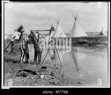 Deux Amérindiens, portant des coiffes de plumes, observent du matériel photographique près d'un ruisseau avec des tipis en arrière-plan. Cette image capture leur interaction avec les premières technologies photographiques. Banque D'Images