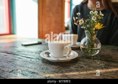 Deux tasses de café et d'un vase de fleurs sur une table de bois, la jeune fille tient dans sa main une tasse de café dans l'arrière-plan. Une photo indique une réunion de personnes et d'un passe-temps commun. Banque D'Images