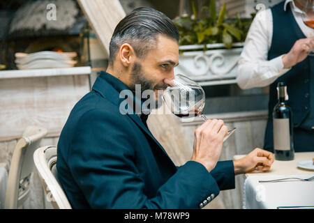 Bel homme riche barbu élégant avec moustache et barbe holding glass et renifler l'odeur de vin rouge au restautant de dégustation. Banque D'Images