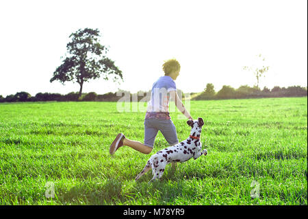 Un garçon court à travers un champ de soleil avec un chien dalmatien en essayant de garder en place. Banque D'Images