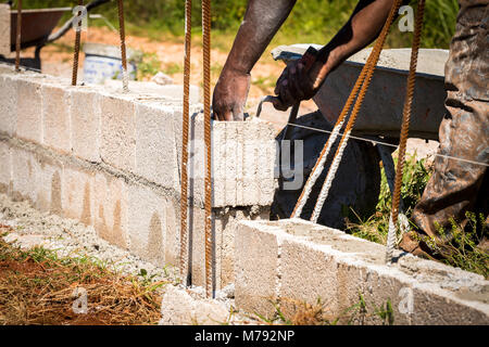 Travailleur de la construction sur site à l'aide d'une truelle pour répandre sa brouette de ciment humide entre les blocs de béton pour maintenir en place les blocs sur des tiges d'acier Banque D'Images