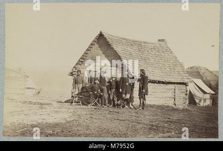 Une photographie historique montrant un groupe de soldats et deux jeunes hommes, dont un afro-américain, debout à l'extérieur des quartiers de cabanes en rondins, capturant un moment pendant la guerre de Sécession. Banque D'Images
