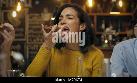 Beautiful Hispanic Woman mange Pizza Slice à l'élégant restaurant. Elle est entourée de ses amis. Banque D'Images