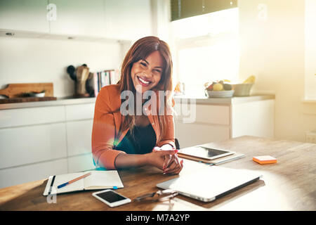 Souriante jeune femme entrepreneur africain assis à sa table de cuisine tout en travaillant de chez vous. Banque D'Images