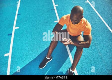 Jeune homme africain runner siégeant seul sur les voies d'une piste de course contrôle de son temps sur sa montre sur une journée ensoleillée Banque D'Images