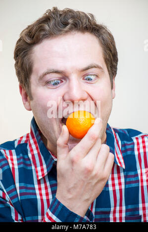 Portrait of young Caucasian man l'origine ethnique, la bouche ouverte de mandarine holding dans la main droite et en essayant d'avaler tout entière. Banque D'Images