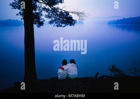 Deux filles préadolescentes siéger ensemble hugging regardant le coucher du soleil sur un lac dans le Vermont, USA. Banque D'Images