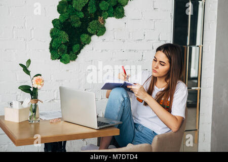 Une jeune fille est un indépendant assis dans un café avec un ordinateur portable et écrit dans un carnet de notes Banque D'Images